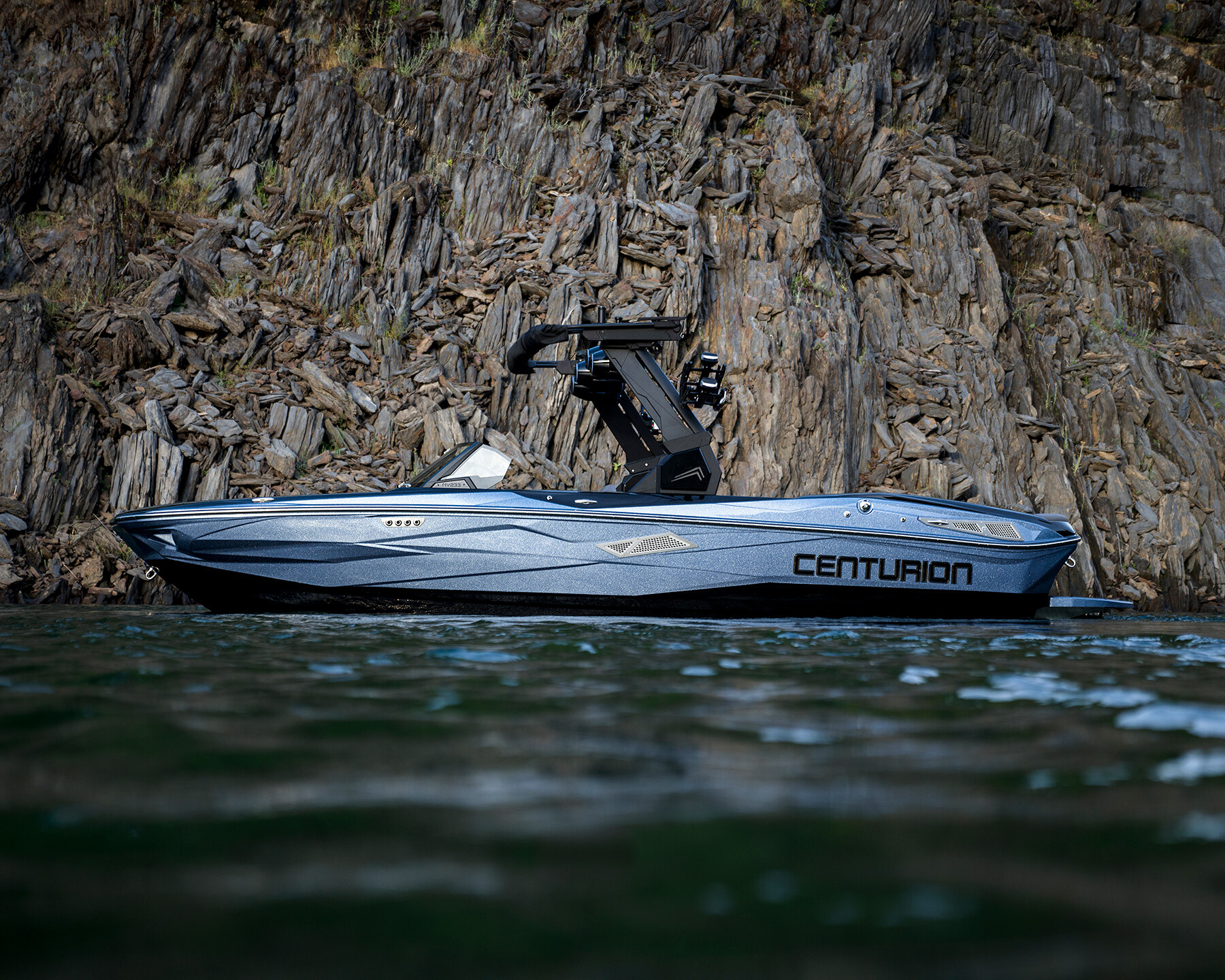 A metallic blue Centurion Nv233 speedboat floats on water near a rocky shoreline with steep, layered cliffs in the background.