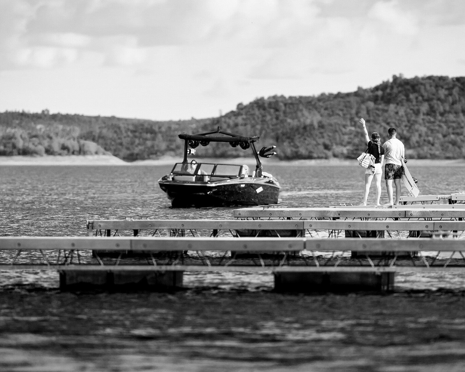 A Centurion Nv233 approaches a dock where two people stand, one raising an arm, with wooded hills and water visible in the background.