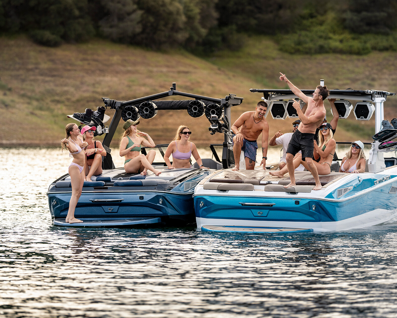 Two motorboats, including a Centurion Nv233, are side by side on a lake. Several people in swimwear relax, chat, and take photos on the Nv233 Wake Boat, while one person stands and points toward the sky.