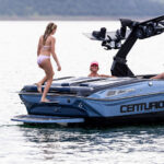 A woman in a pink bikini steps onto a blue Centurion Nv233 wake boat where two people are seated, one wearing a pink cap, on a calm body of water.