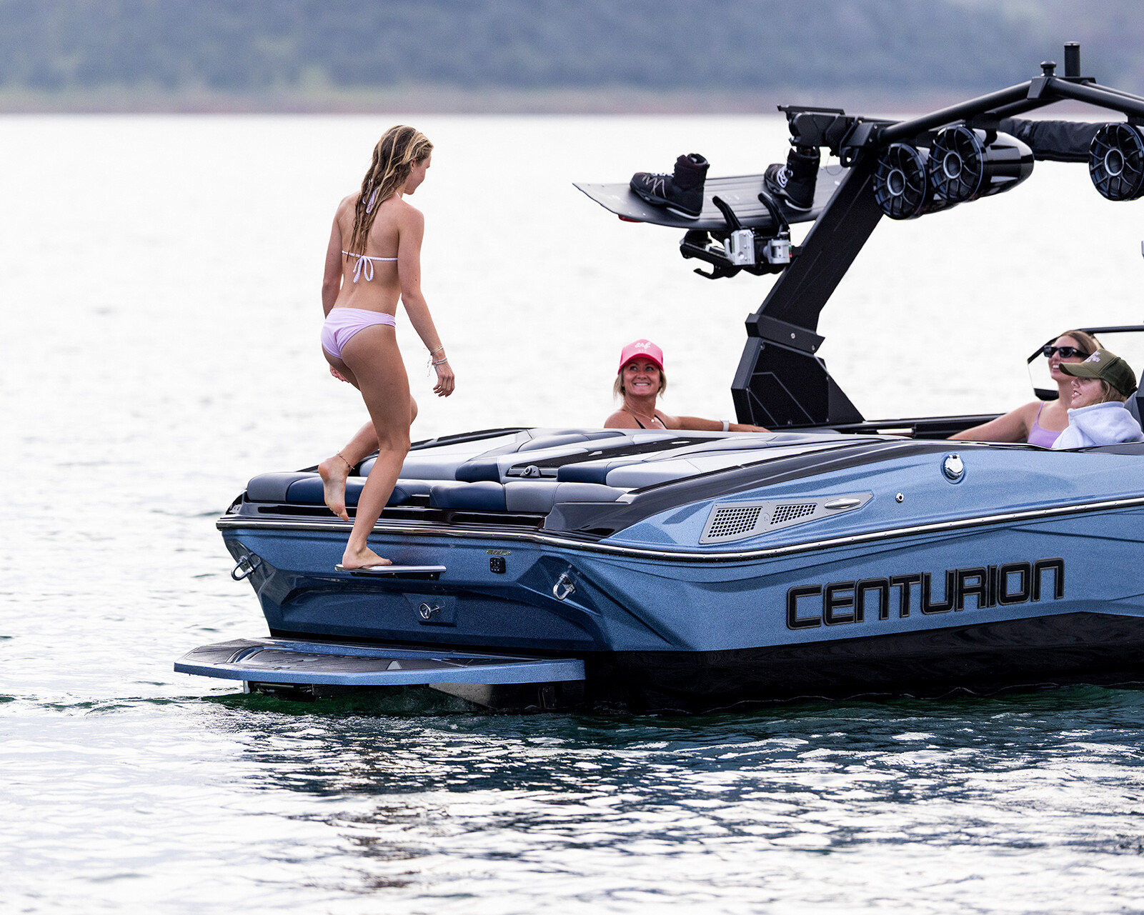 A woman in a pink bikini steps onto a blue Centurion Nv233 wake boat where two people are seated, one wearing a pink cap, on a calm body of water.