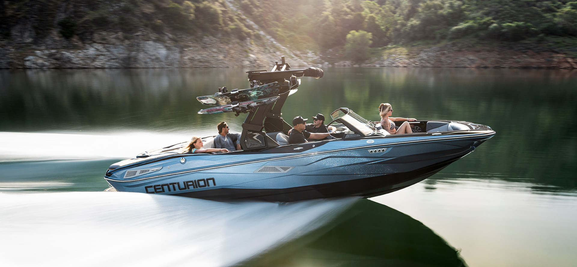 A blue Centurion Nv233 speedboat with four people onboard moves quickly across a calm lake, surrounded by trees and a rocky shoreline.