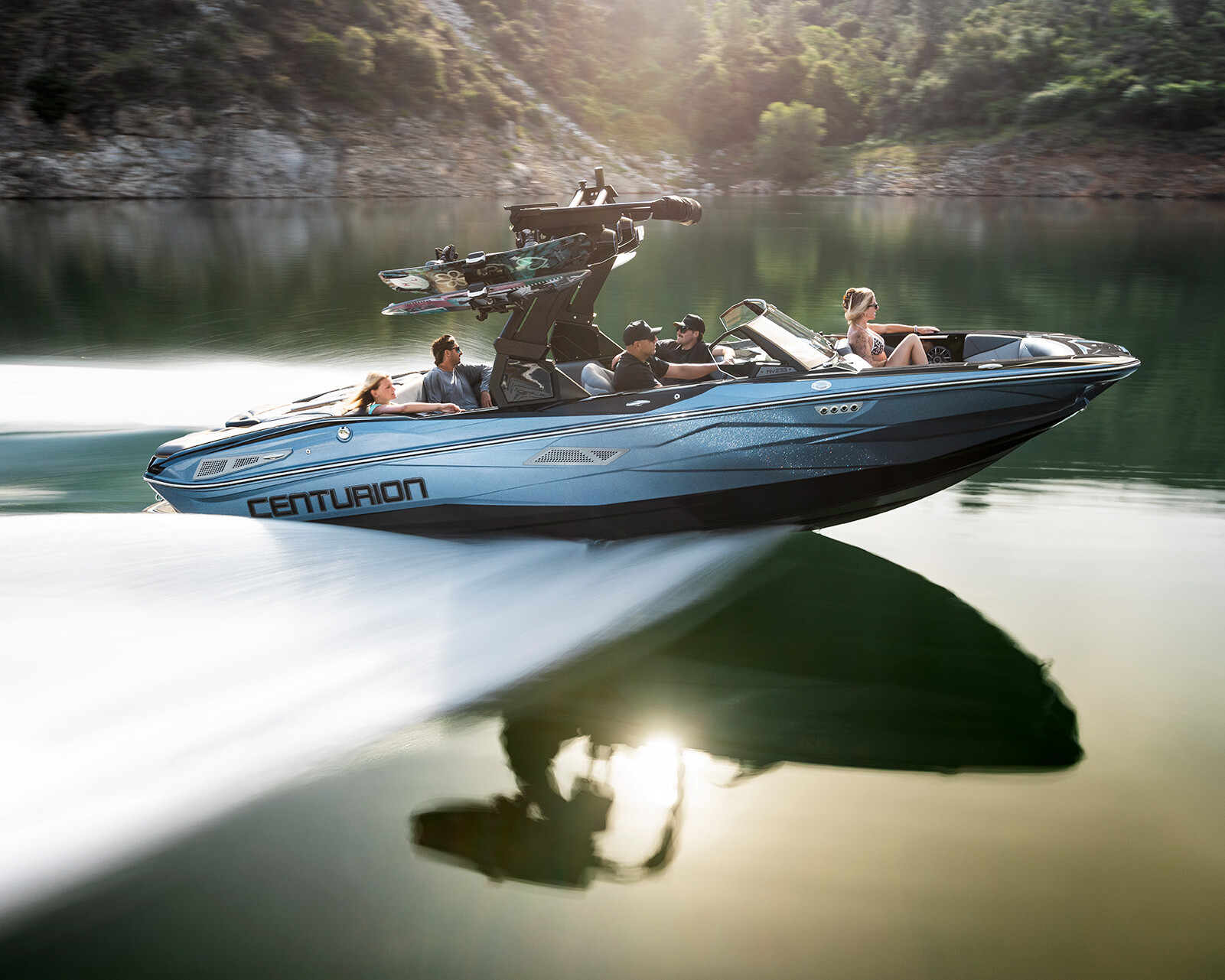 A Centurion Nv233 speedboat with four people moves quickly across a calm lake, with wakeboards mounted on top and trees visible in the background.