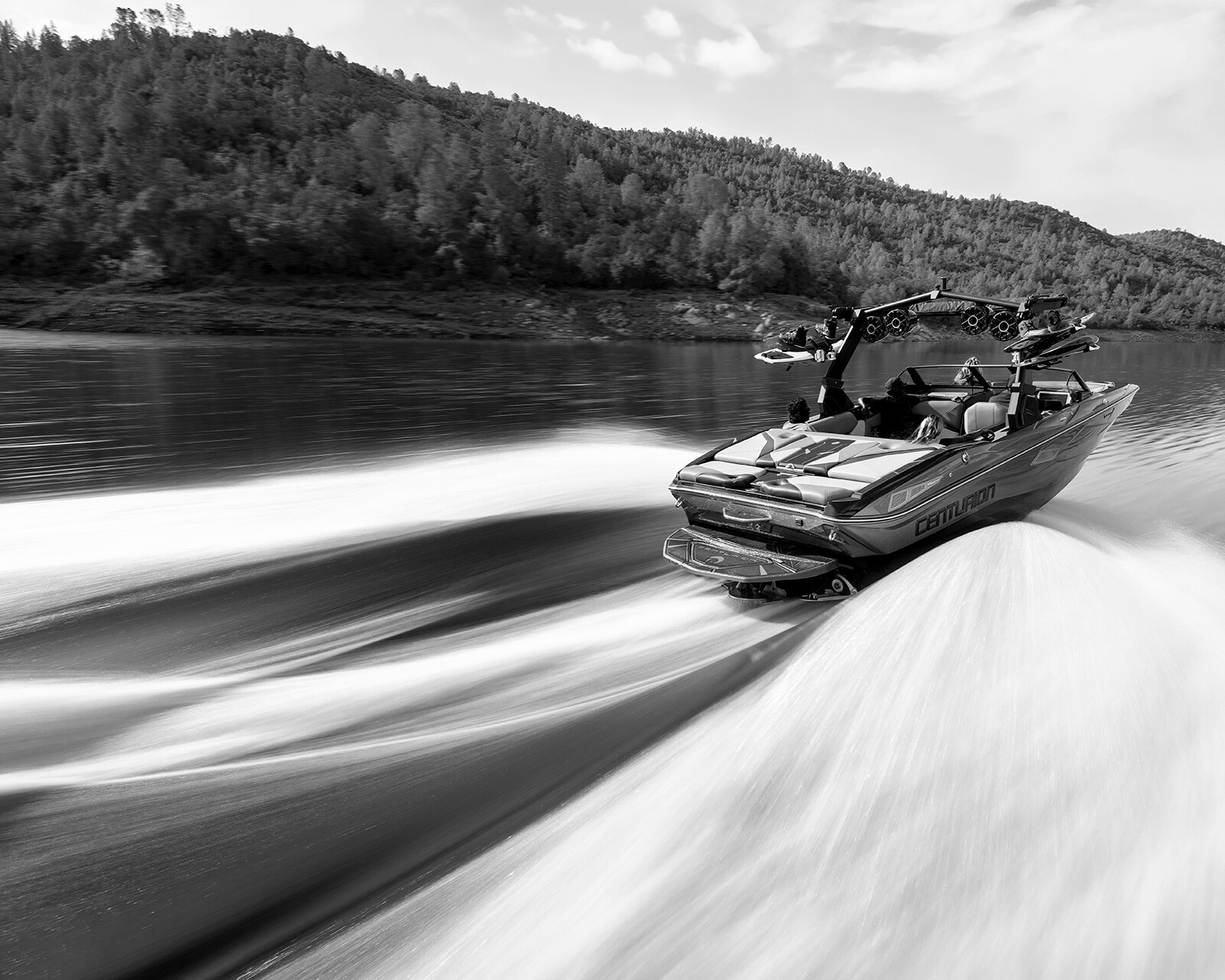 A Centurion Nv233 speedboat moves quickly across a calm lake, leaving a large wake behind, with forested hills visible in the background.
