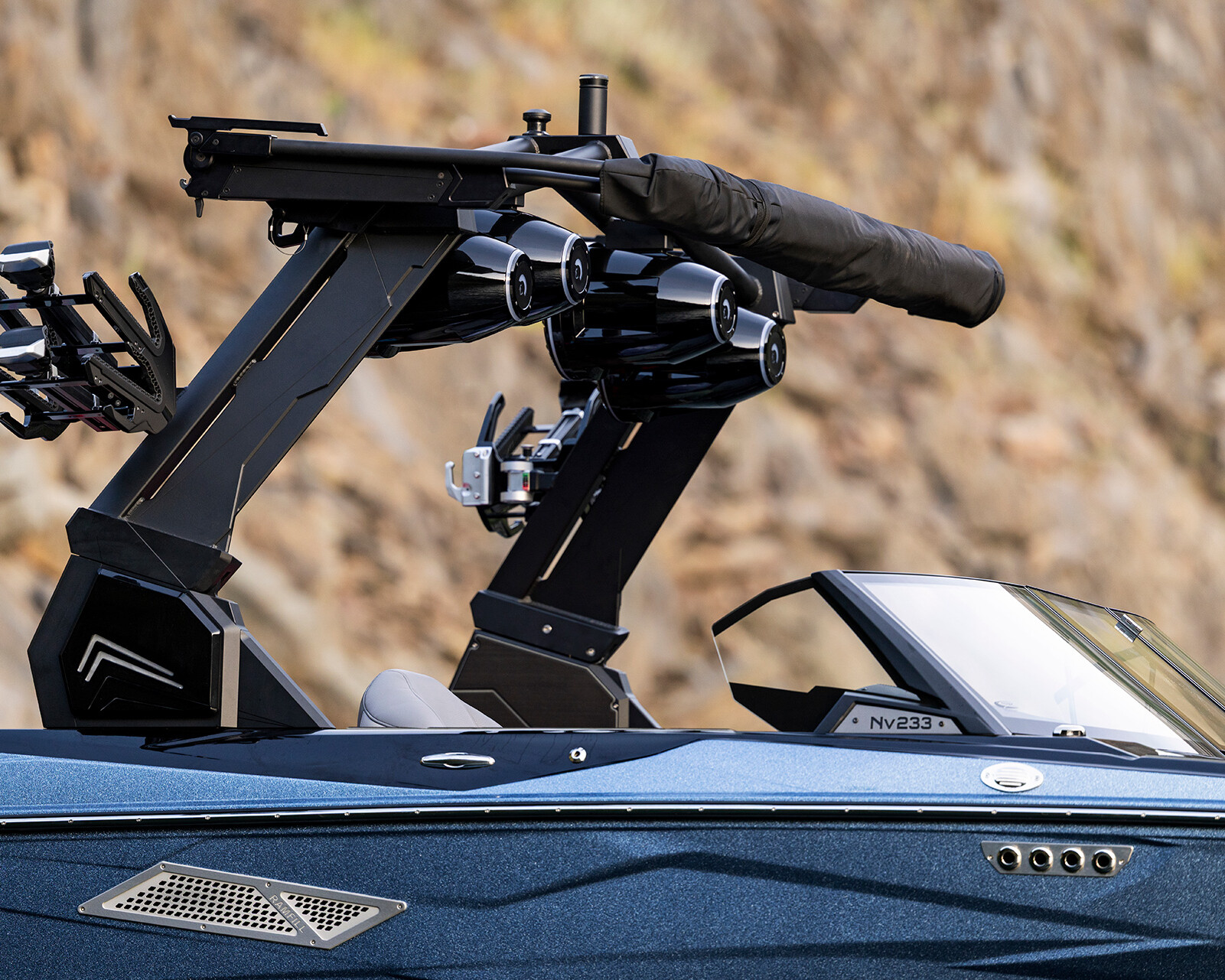 Close-up of a blue Nv233 Wake Boat with two black wakeboard racks, mounted speakers, and a folded bimini top, set against a rocky background.