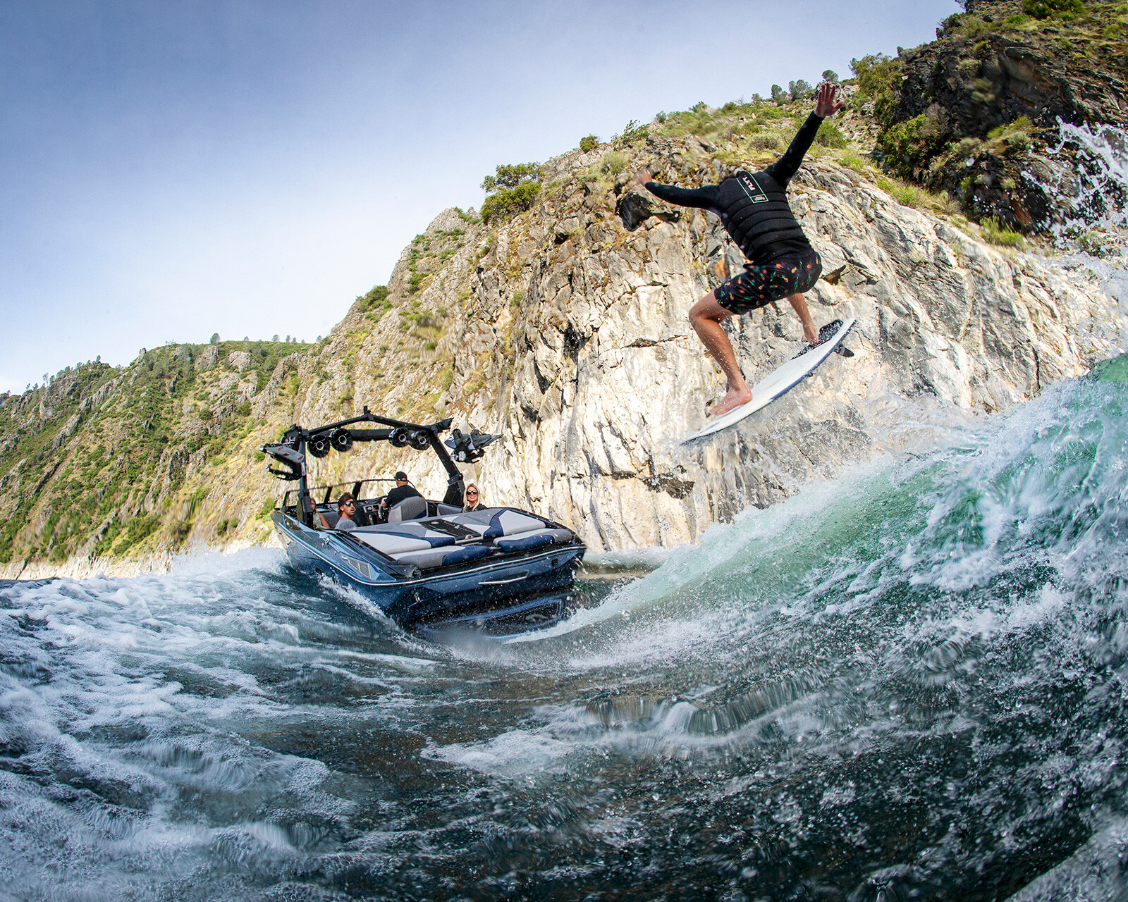 A person wakeboarding behind a Centurion Nv233 wake boat performs an airborne trick on a river near rocky cliffs under a clear sky.