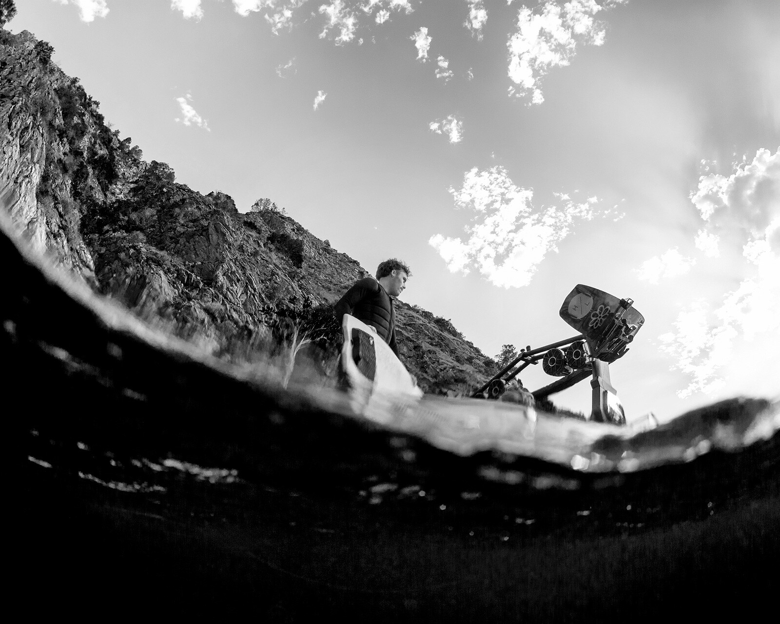 A person stands near a rocky shore operating a camera on a tripod, photographed from a low angle partially submerged in water, with an Nv233 Surf Boat visible in the background, all captured in striking black and white.