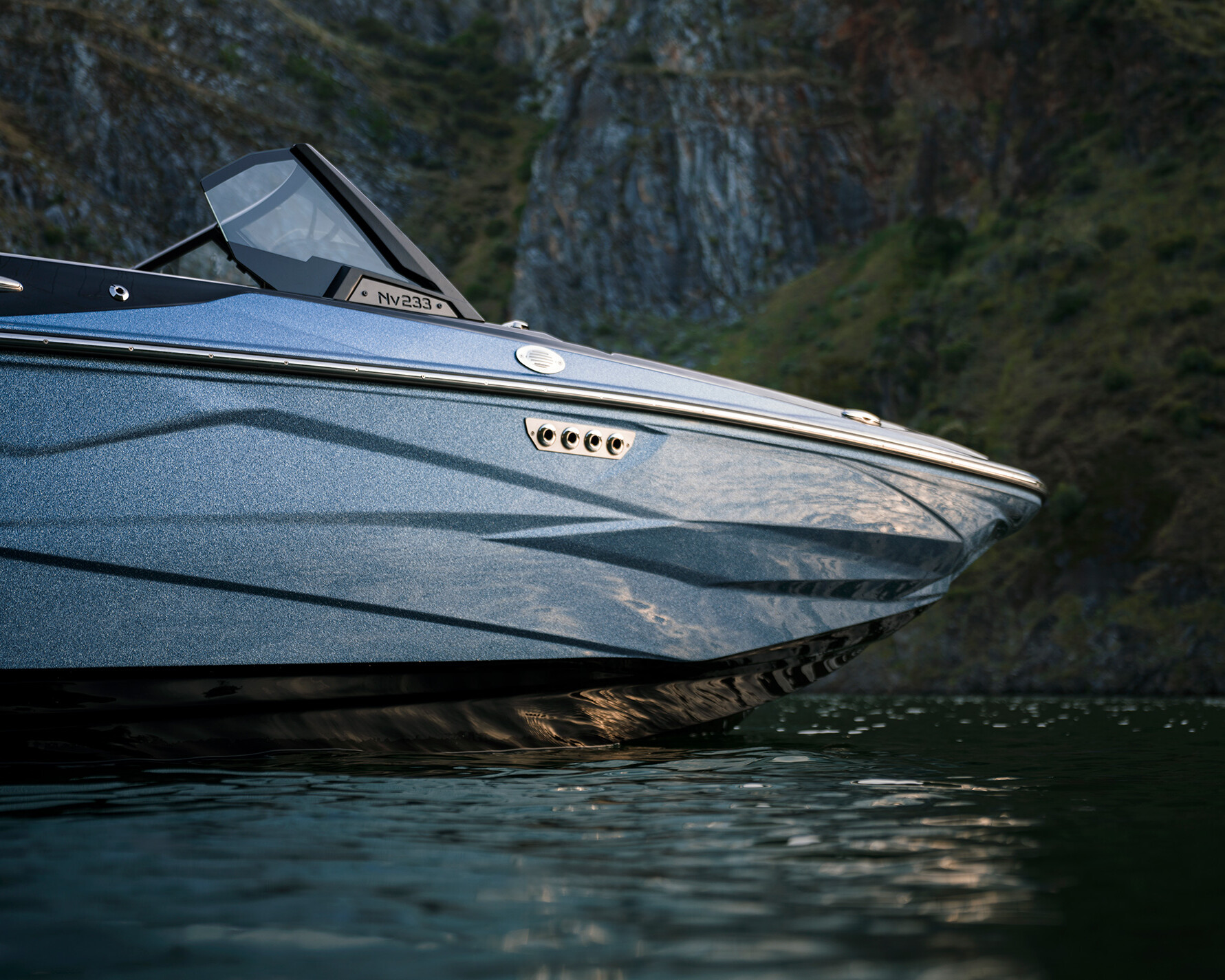 Close-up of the front section of a sleek, modern Centurion Nv233 surf boat floating on calm water with rocky cliffs in the background.