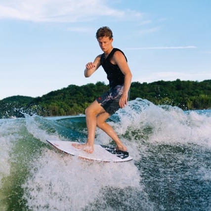 A person wearing a black tank top and shorts is surfing on a wave, with greenery and hills in the background.