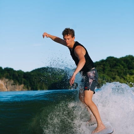 Person surfing on a wave with greenery in the background under a clear blue sky.