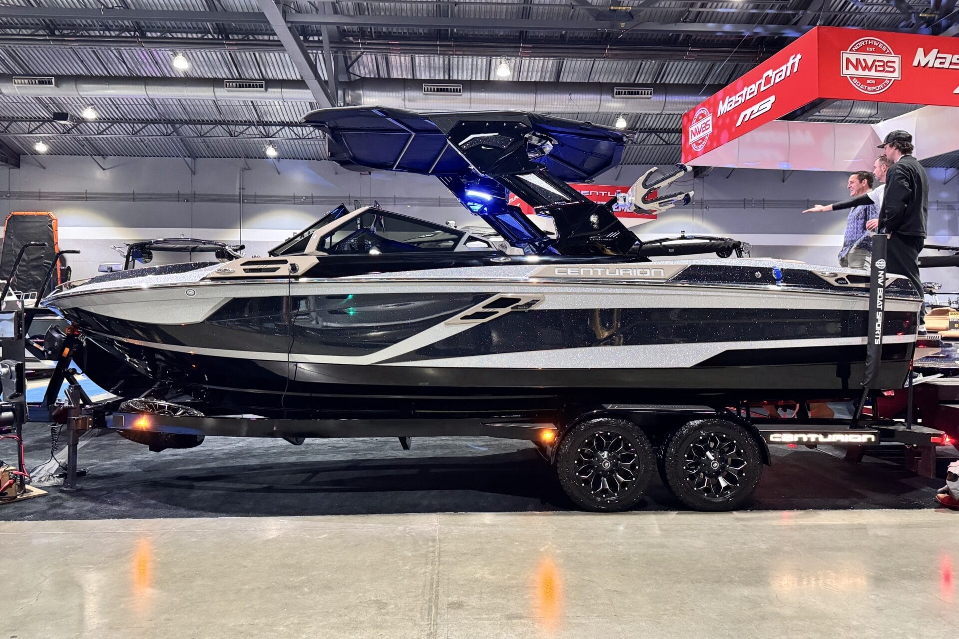 A sleek black and silver Centurion boat on display indoors, mounted on a trailer with shiny black wheels.