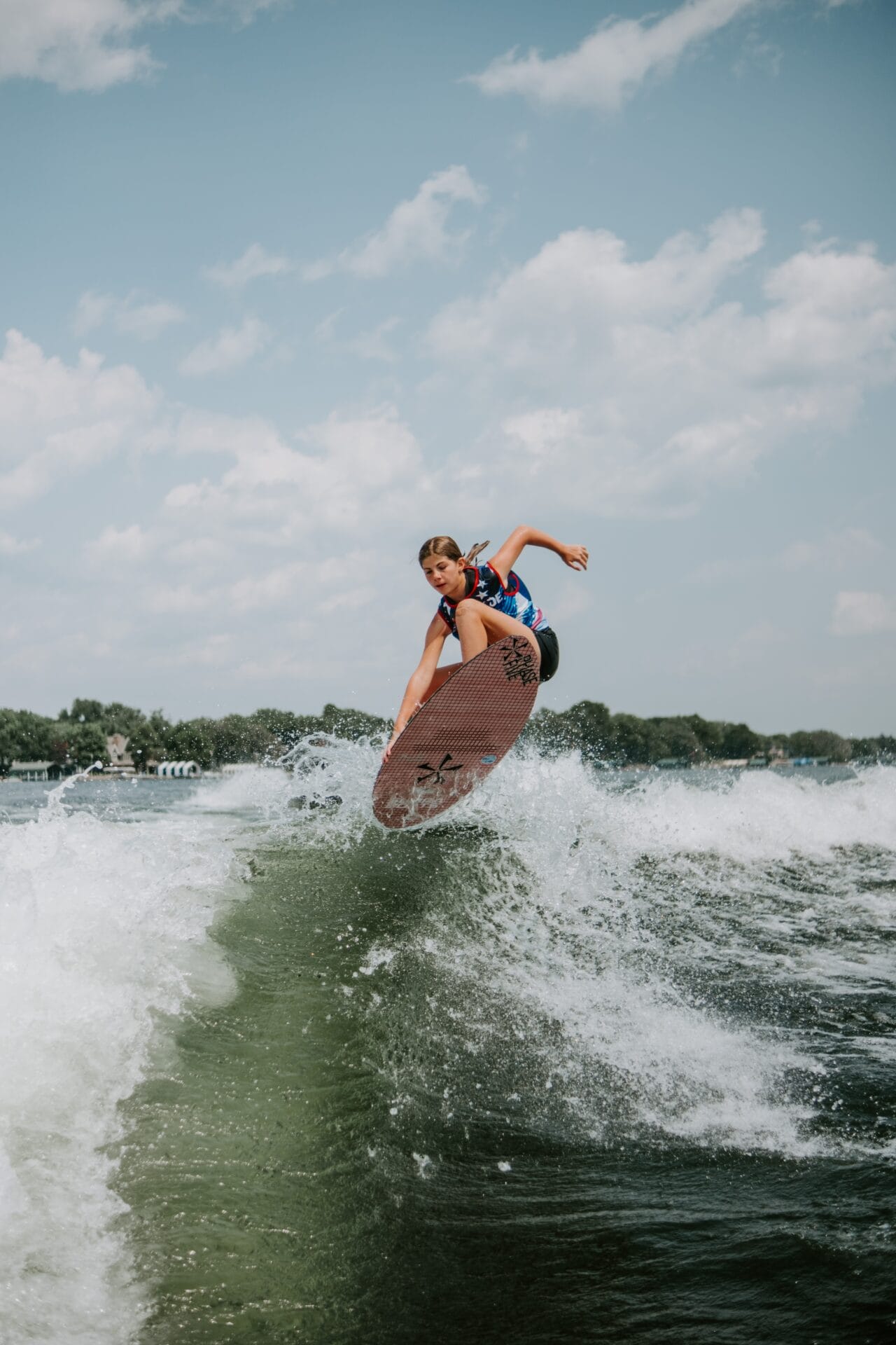 Person surfing on a wave under a partly cloudy sky, performing a jump on a red surfboard with a vibrant landscape in the background.