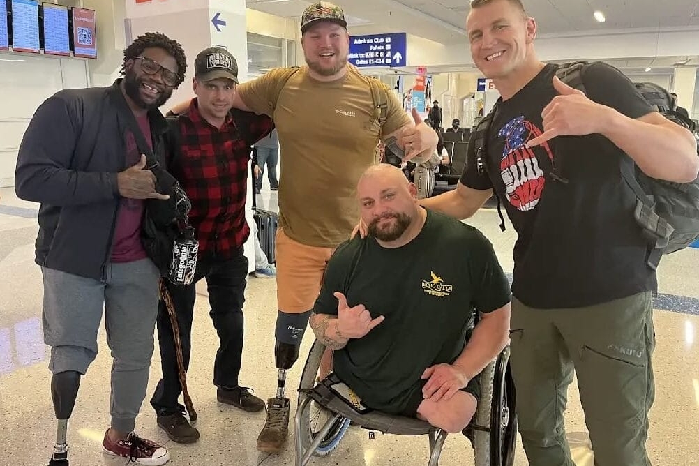 A group of five men, some with prosthetic legs, pose and smile at an airport terminal. They are casually dressed and making hand gestures. Airport signage is visible in the background.