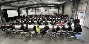 A large group of people seated in a warehouse setting, facing a presentation screen with "100" displayed.