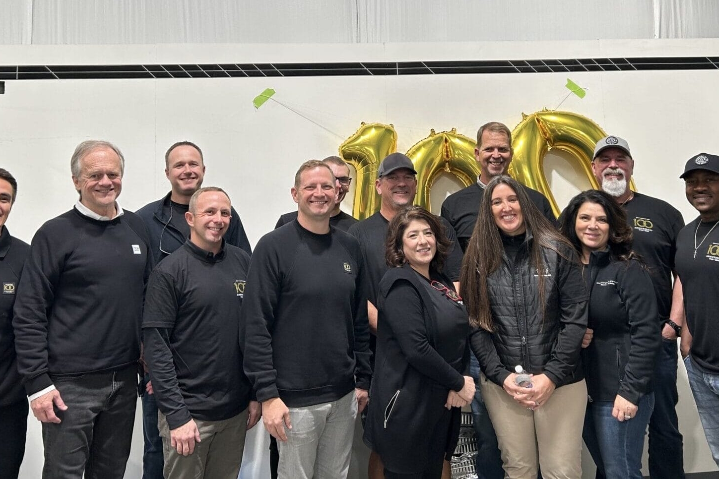 A group of thirteen people poses indoors, smiling in front of golden balloon numbers 