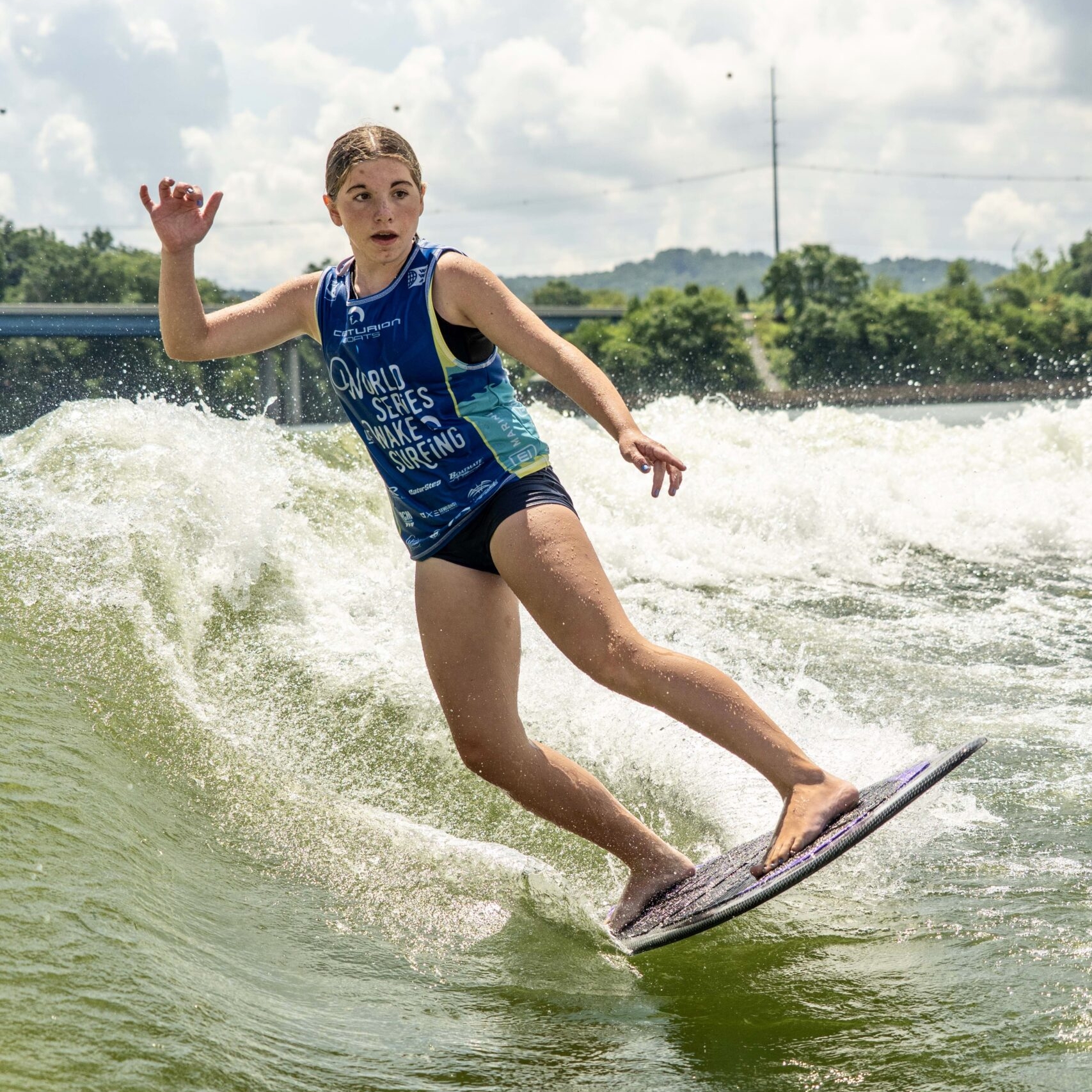 Aria Hellemn, in a blue tank top and black shorts, surfs on a wave under a partly cloudy sky, maintaining balance with one arm raised.