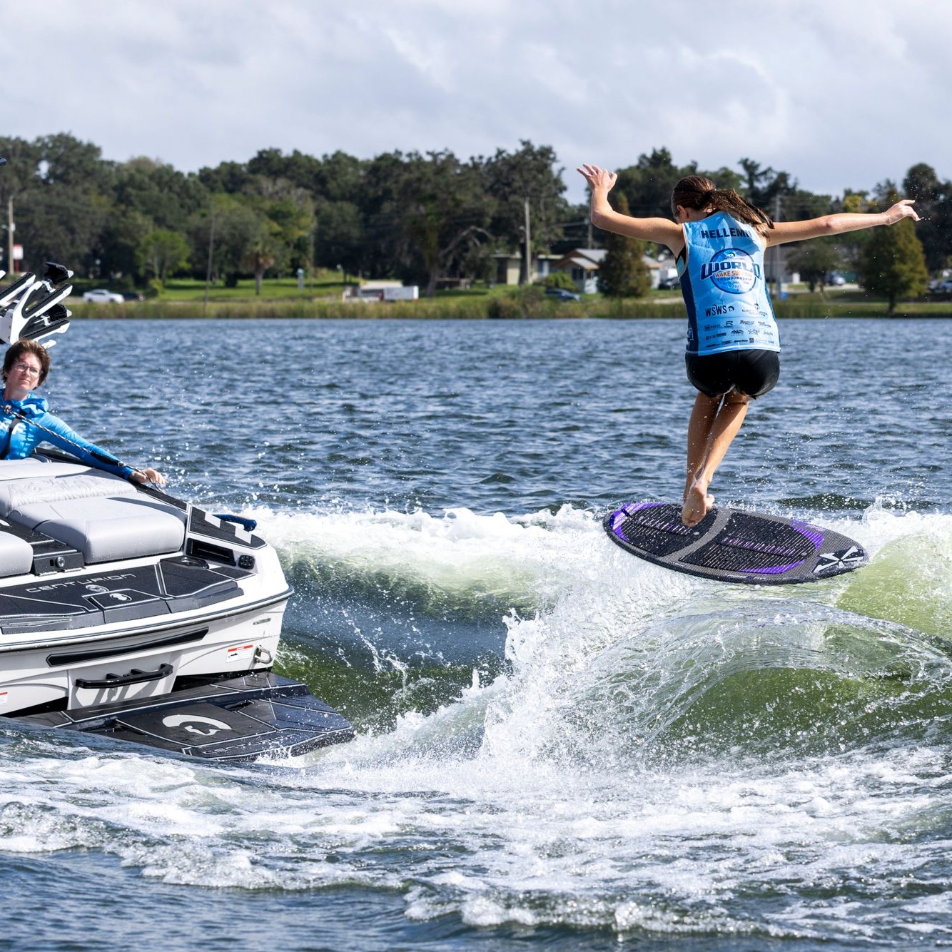 A person surfs on a wave behind a motorboat, while several people watch and take photos from inside the boat.
