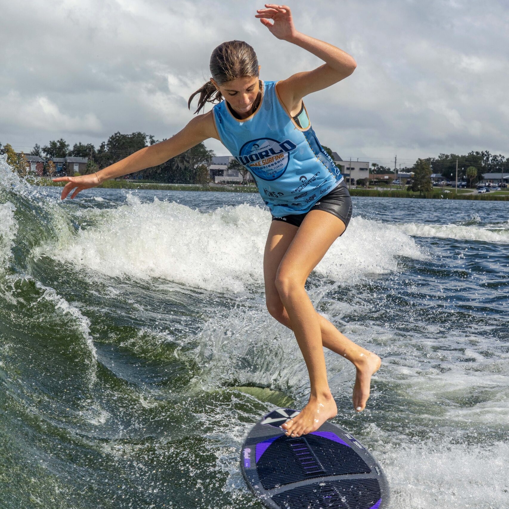 A young woman wearing a blue top and shorts is riding a skimboard on a wave, balancing with arms outstretched, with water splashing around her under a partly cloudy sky.