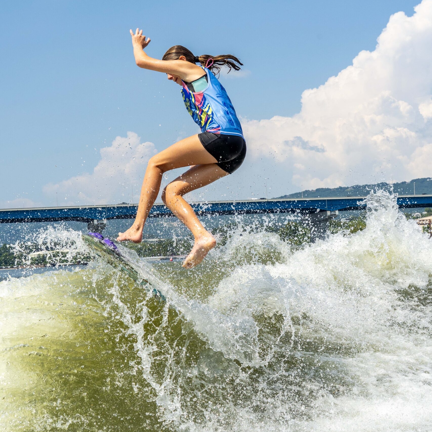 A person wearing a blue shirt and black shorts is jumping on a surfboard above a wave, with water splashing around and a blue sky in the background.
