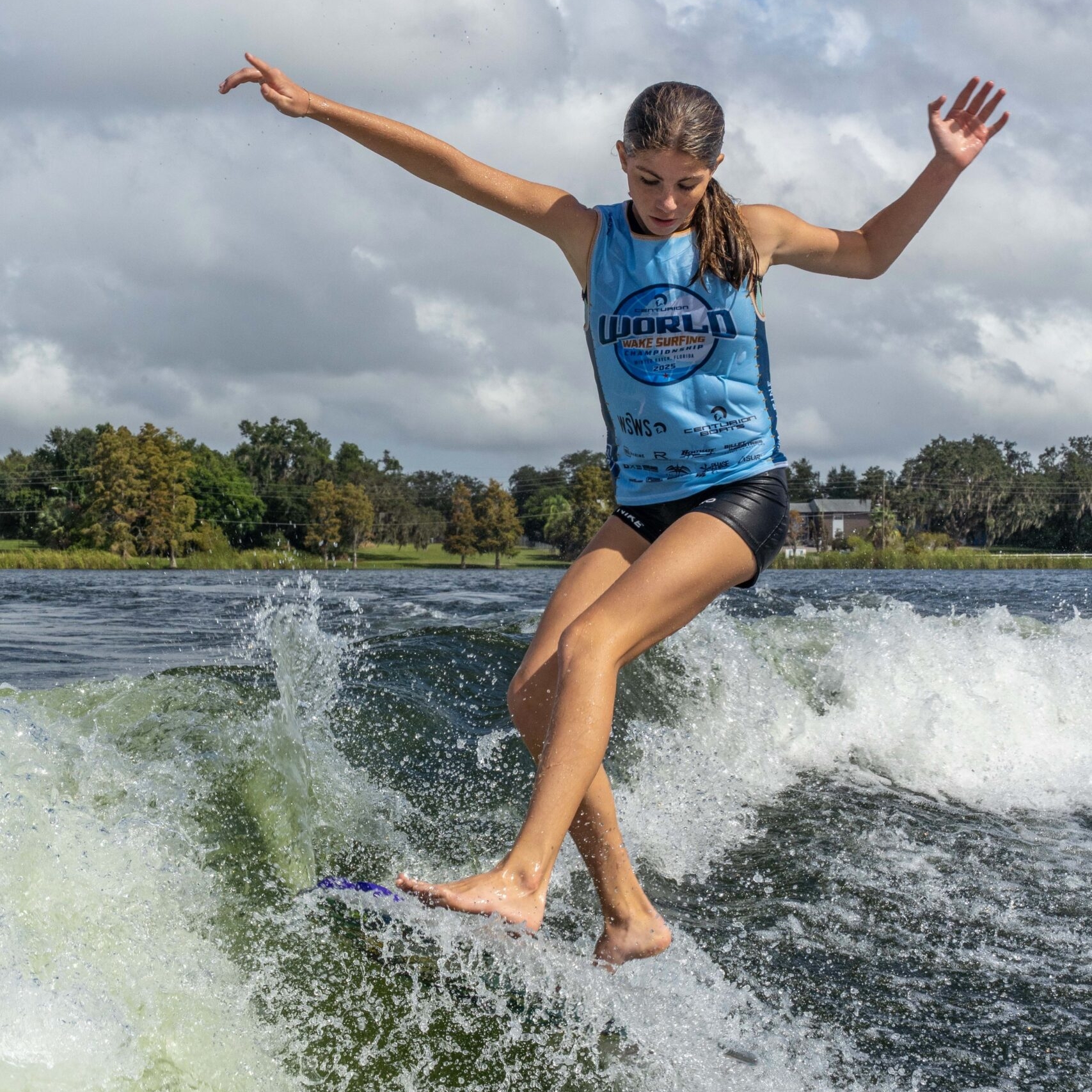 A young woman in a blue tank top and shorts rides a wave on a surfboard with arms outstretched for balance under a partly cloudy sky.