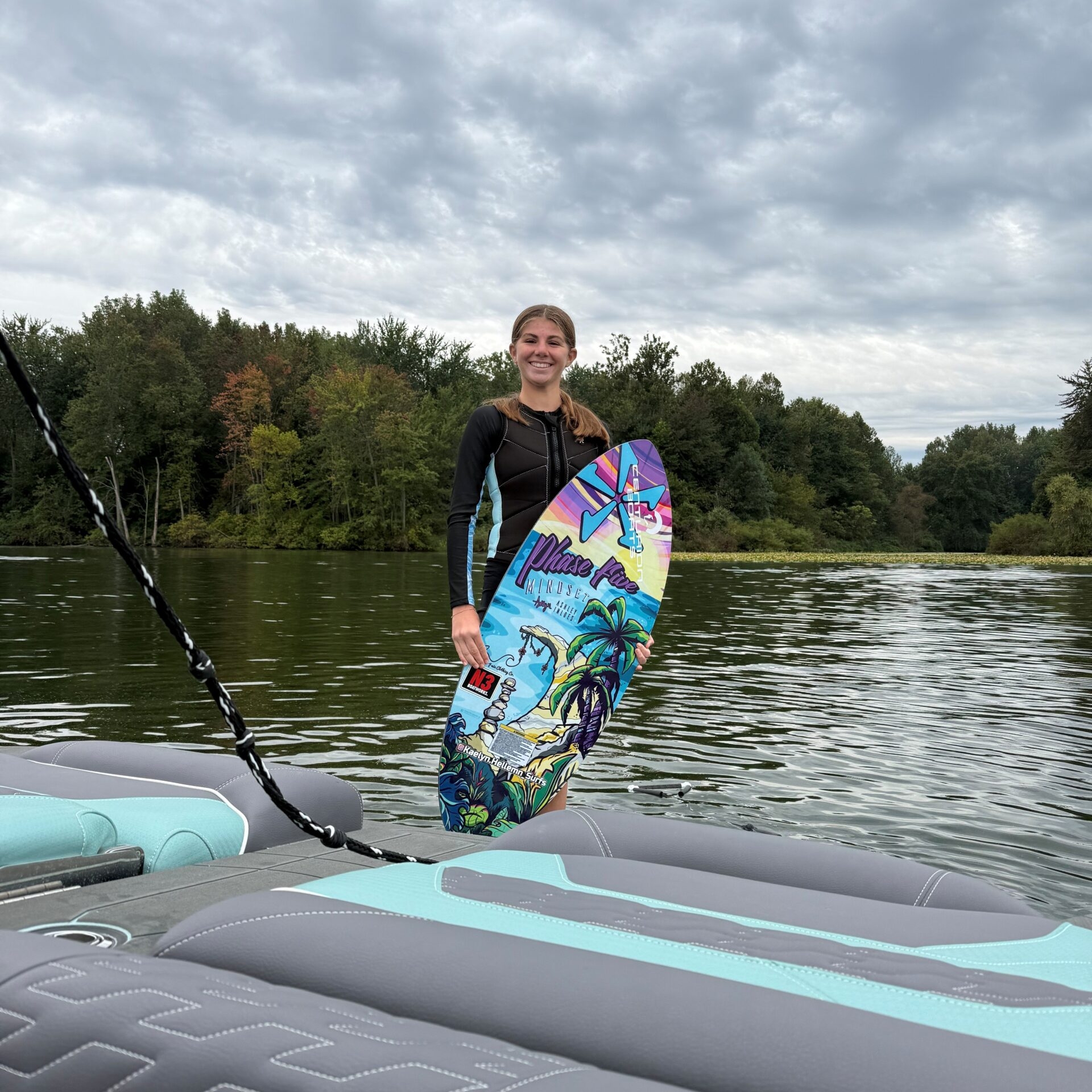 A person in a wetsuit stands on a boat holding a colorful wakeboard, with a lake and trees in the background under a cloudy sky.