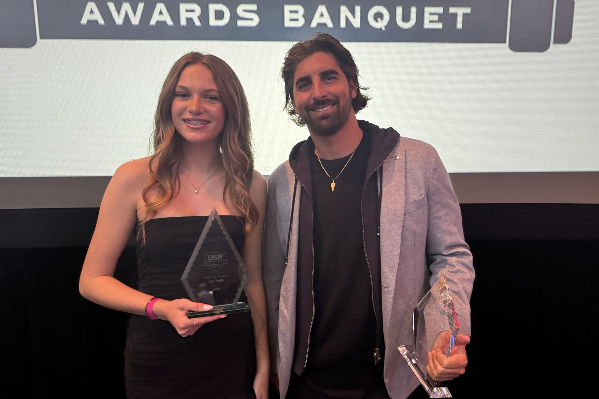 Two people holding awards and smiling at a banquet, standing in front of a backdrop that reads 