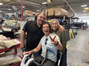 Three people inside a warehouse, two standing and one seated on a scooter, smiling and posing for the camera. The seated person is making a peace sign with their hand.