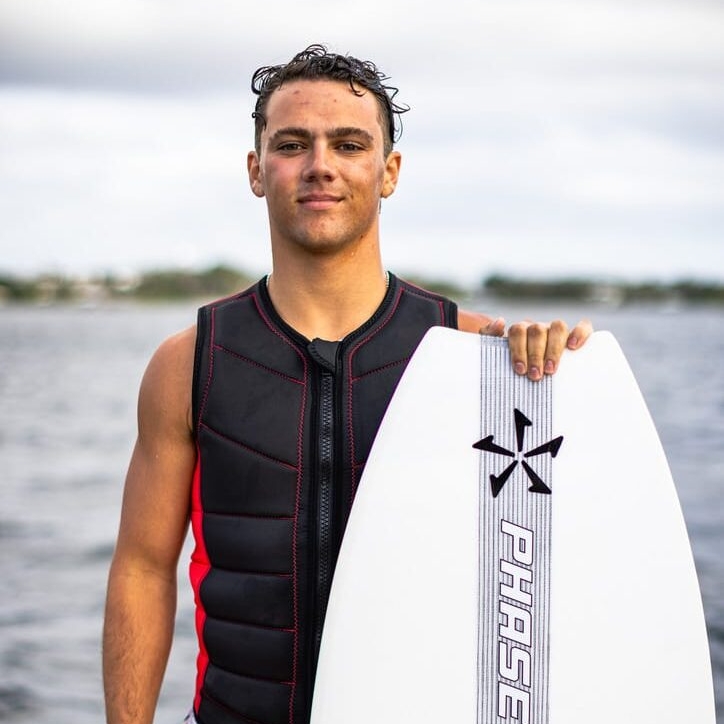 Sammy Goncalves stands by the water, holding a white surfboard vertically. Wearing a black protective vest and patterned shorts, they gaze at the overcast sky, ready for the next wave.