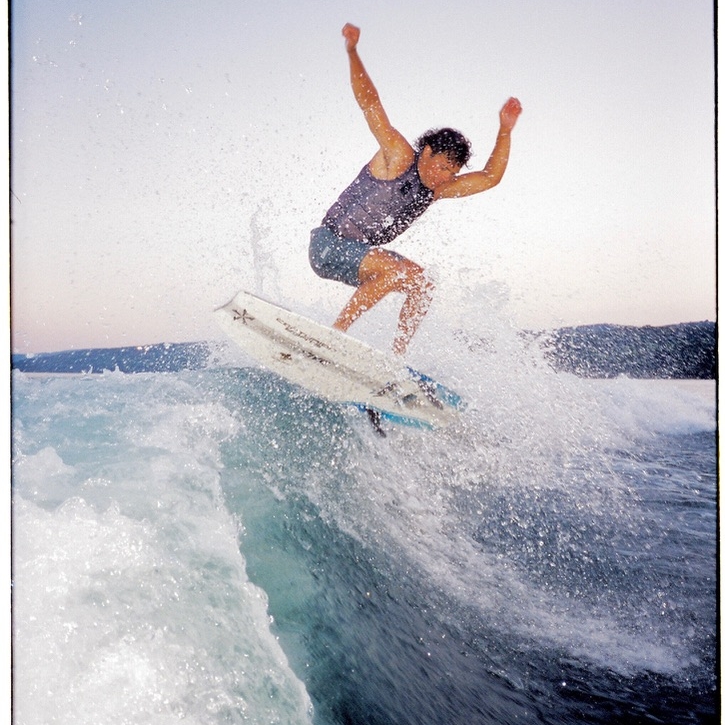 Sammy Goncalves rides a wave on a surfboard, mid-air above the water, with arms raised and ocean spray around them.
