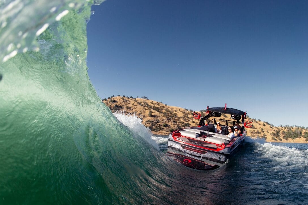 A speedboat with passengers navigates through a wave on a sunny day with hills in the background.