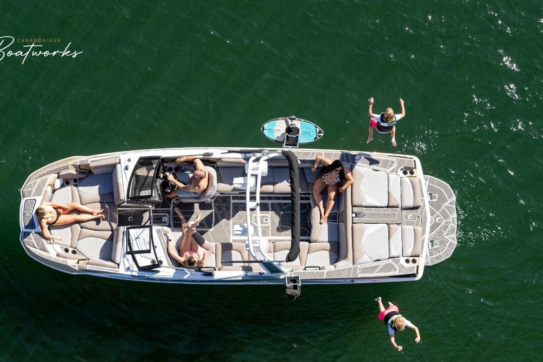 Aerial view of a boat on the water with four people. Two are seated inside, one is standing at the rear, and one person is jumping into the water.