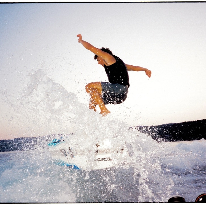 Sammy Goncalves, in a black tank top and shorts, jumps off a wakeboard, splashing water as the sun sets over the lake and hills in the background.