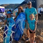 A family of four poses on a sandy beach. They are holding two surfboards and wearing matching blue shirts. A tent and chairs are in the background.