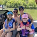 A family of four on a boat: two adults and two girls wearing life vests and caps, smiling at the camera. Water and greenery are visible in the background.