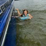 A person in a blue life vest smiles while holding onto the side of a blue boat, floating on a body of water.