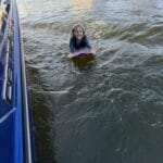 Child smiling while riding a boogie board on water next to a blue boat.