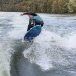 A person surfs on a river, performing a jump. The background shows a wooded hillside with autumn-colored leaves.