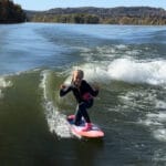 A person in a wetsuit rides a wave on a pink surfboard, making hand gestures, with a scenic view of trees and hills in the background.