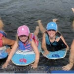 Four people in life vests and caps lie on surfboards in the water, smiling at the camera.