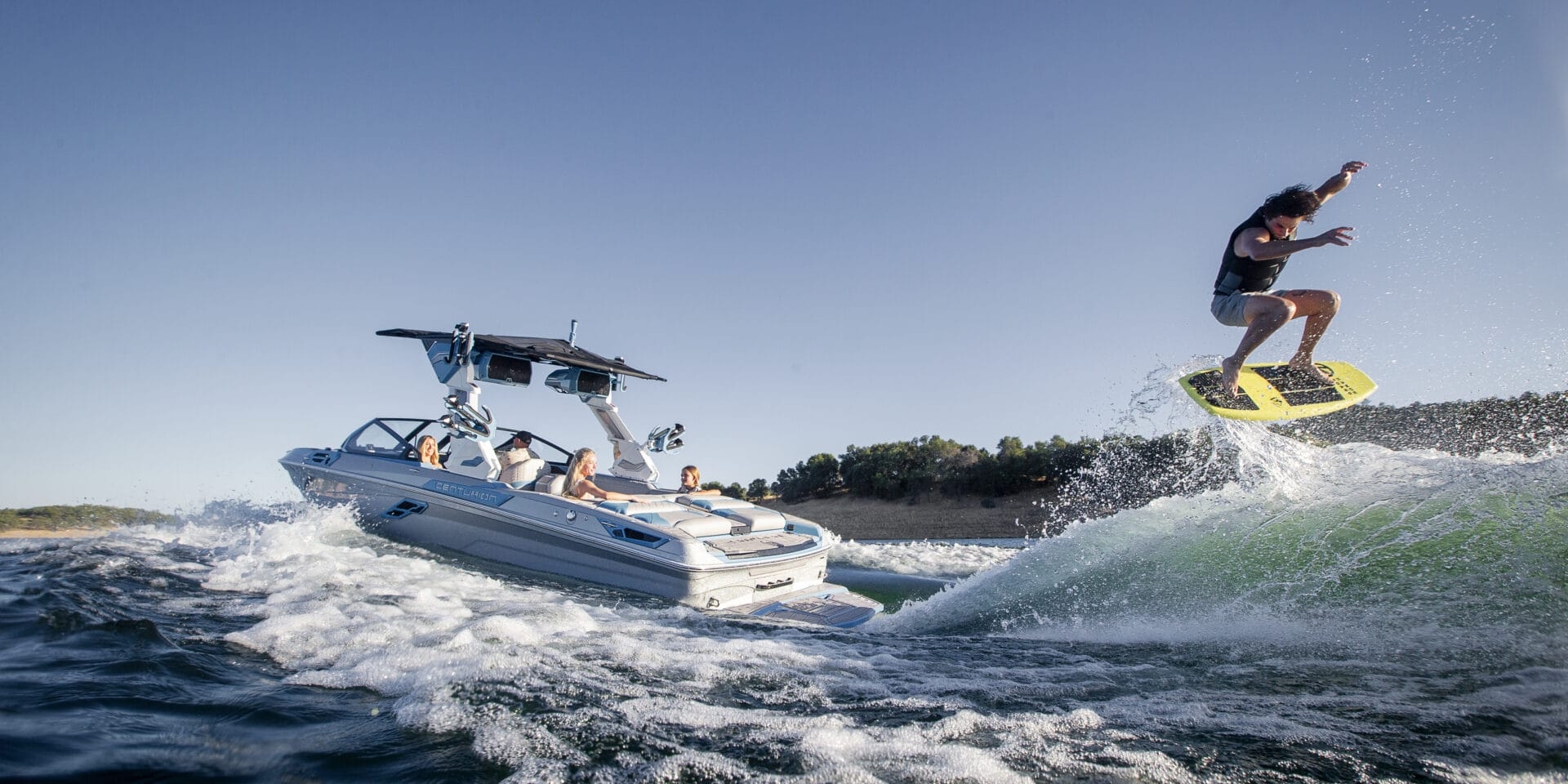 A person is wakesurfing and performing a jump behind a motorboat on a sunny day, with several people riding in the boat.