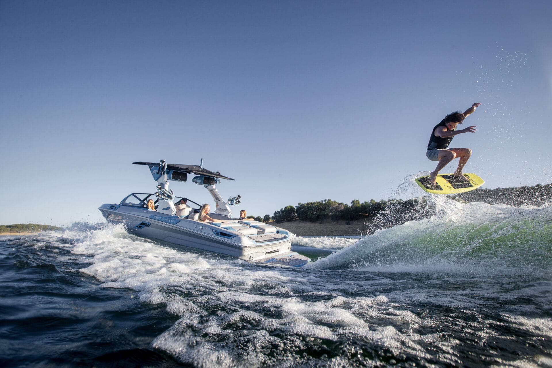 A person is wakesurfing and performing a jump behind a motorboat on a sunny day, with several people riding in the boat.