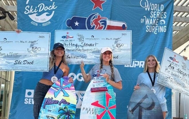 Three women stand on a podium holding wakeboards and oversized prize checks at a wake surfing competition, with event banners in the background.
