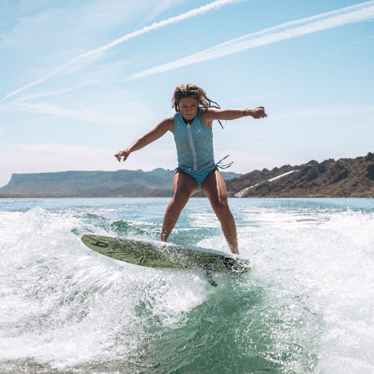 A person in a blue life vest surfs on a wave in open water with hills and blue sky in the background.