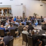 A group of people sit at round tables in a large room, listening to a speaker presenting at the front near a projection screen.