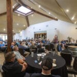 A group of people seated at round tables listen to a speaker presenting at the front of a spacious, well-lit conference room.