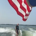 A man rides a wakeboard on a lake behind a boat, with a large American flag prominently displayed overhead. Another person in a white cap sits in the foreground.