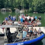 A group of people pose on a dock next to a police boat and a fire boat on a lake, with trees and docks visible in the background.