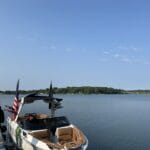A white motorboat with tan seats is docked on a calm lake. An American flag is mounted on the boat, and trees line the distant shoreline under a clear blue sky.