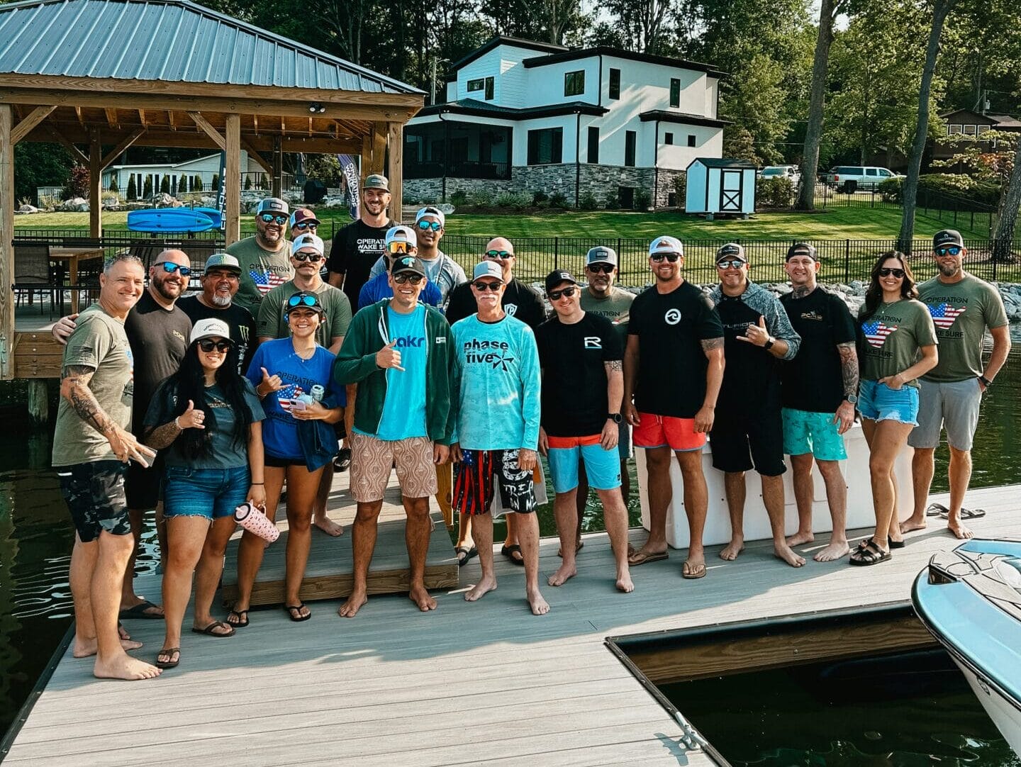 A group of people pose for a photo on a dock by the water, with trees, a gazebo, and a house visible in the background.