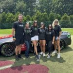 Six people stand in front of a race car on a grassy field, all wearing sunglasses and black t-shirts, with trees and a fence in the background.